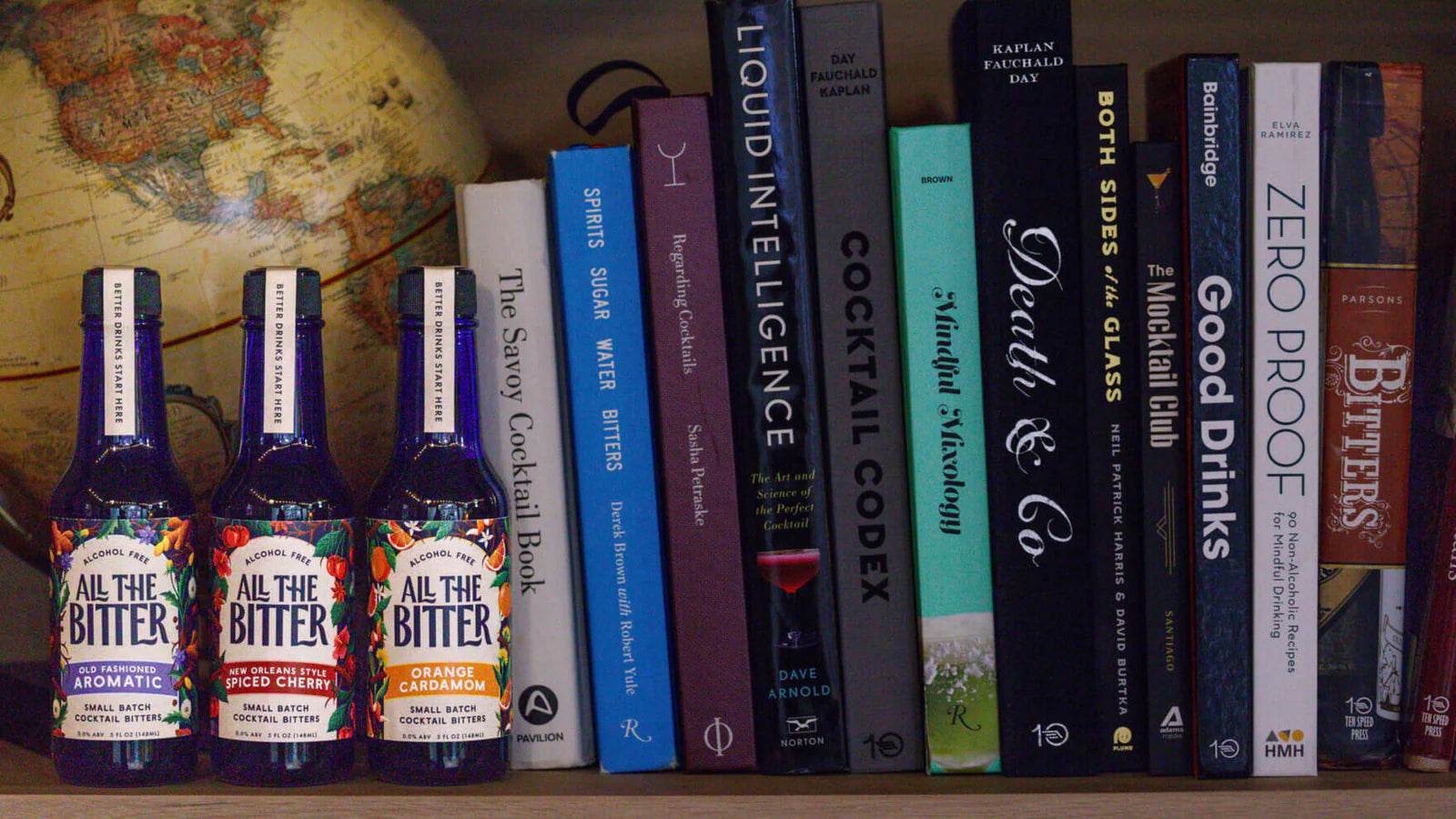 Three bottles of 'All the Bitter' nonalcoholic bitters with cocktail books on a shelf