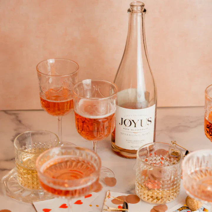 Bottle of JOYUS sparkling wine with glasses filled with the wine on a marble surface.